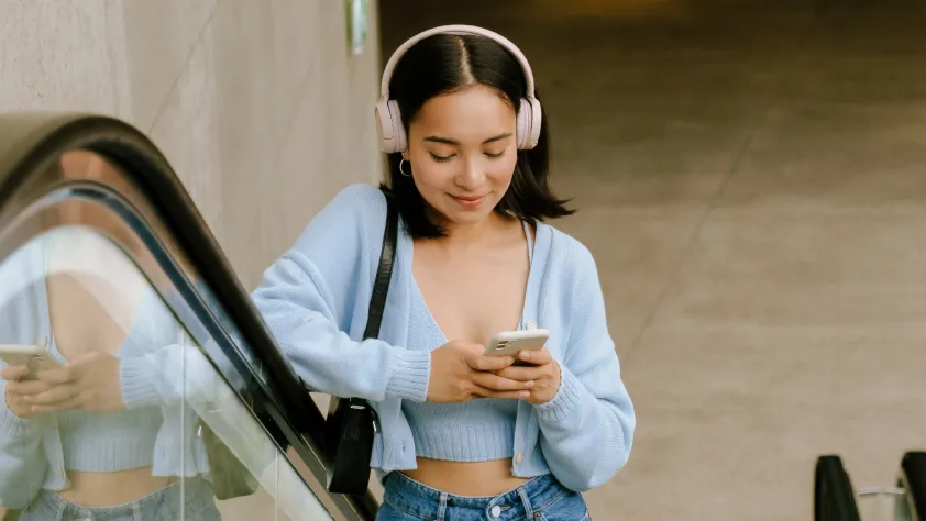 Seseorang sedang menaiki eskalator, mengenakan sweter dan jin biru, serta headphone merah muda. Dia membawa dompet hitam dan membaca pesan bisnis di WhatsApp di ponsel.