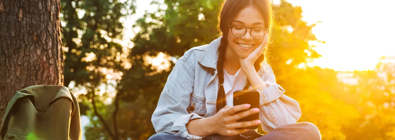 Person sitting in the woods with a sunset in the background. They are wearing a denim jacket, glasses, and jeans, while smiling and looking at phone.