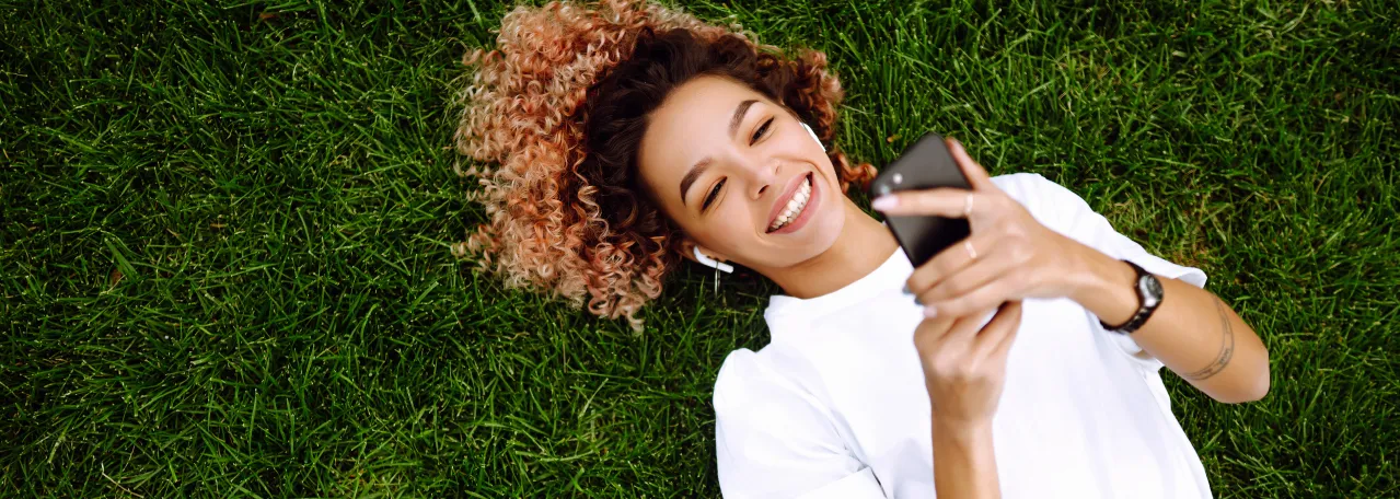 Person laying on the grass and wearing AirPods and a white t-shirt. They are smiling and clicking on an ad that leads to WhatsApp on phone.