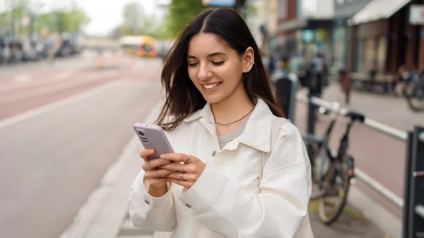 Person standing on Amsterdam street with the city in the background. They are wearing a white jacket while smiling and looking at smartphone.