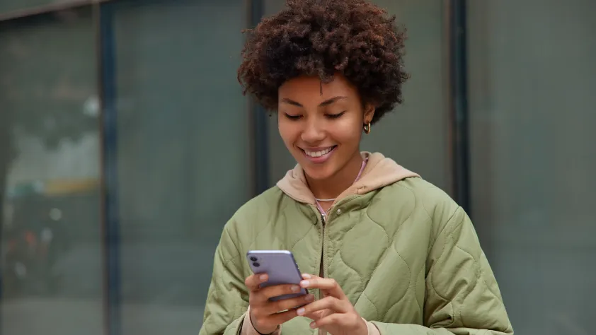Person standing in front of office building windows. They are wearing a light green jacket and smiling while looking at purple iPhone.