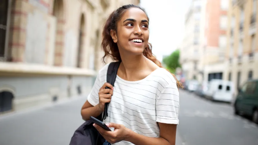 Pessoa caminhando pela rua com uma mochila e vestindo uma camiseta branca listrada. Ela está segurando um celular e sorrindo enquanto olha para trás.