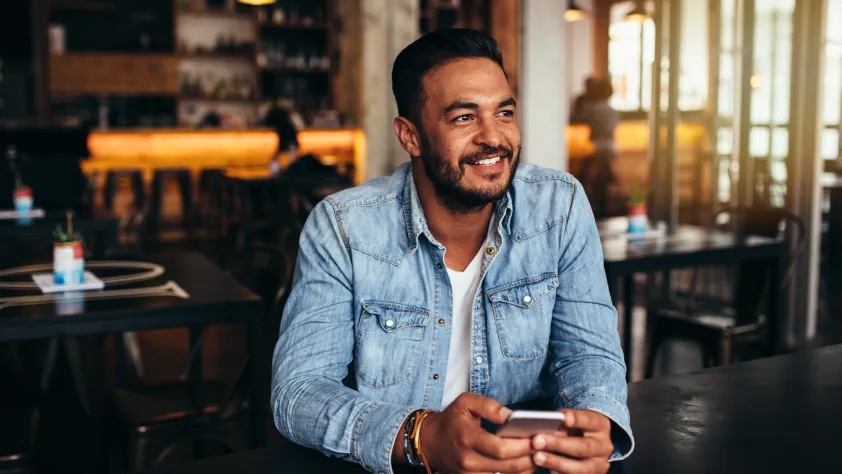 Person sitting in restaurant, wearing a denim shirt and holding phone while looking out the window.