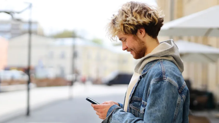 Person standing outside cafe with umbrellas, building, and street in the background. They are wearing a sweatshirt and a jean jacket while looking at a product recommendation via WhatsApp on phone.