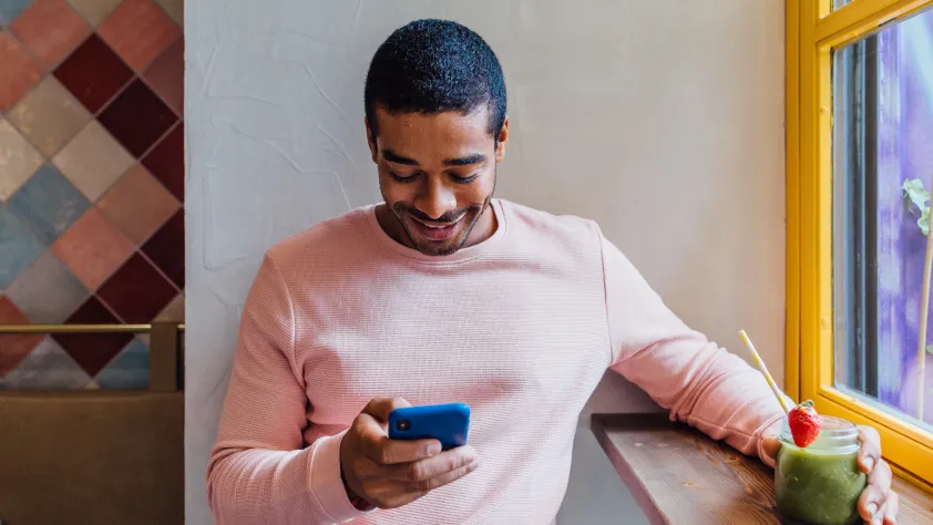 Person sitting in a cafe in front of the window, wearing a pink long sleeve shirt. They are holding onto a matcha latte and smiling at their phone.