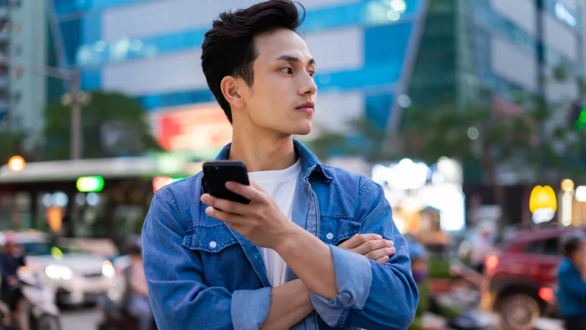 Person standing in front of bustling city background. They are wearing a white t-shirt and a blue denim shirt, while holding a phone and looking off into the distance.