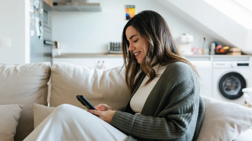 Person sitting on white couch, wearing a white t-shirt and green sweater. They are smiling and looking at an ad on Instagram on their phone.