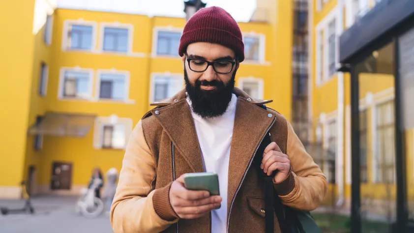 Person walking down the street with yellow buildings in the background. They are wearing a burgundy beanie, glasses, and a brown jacket while reviewing a product catalog on WhatsApp.
