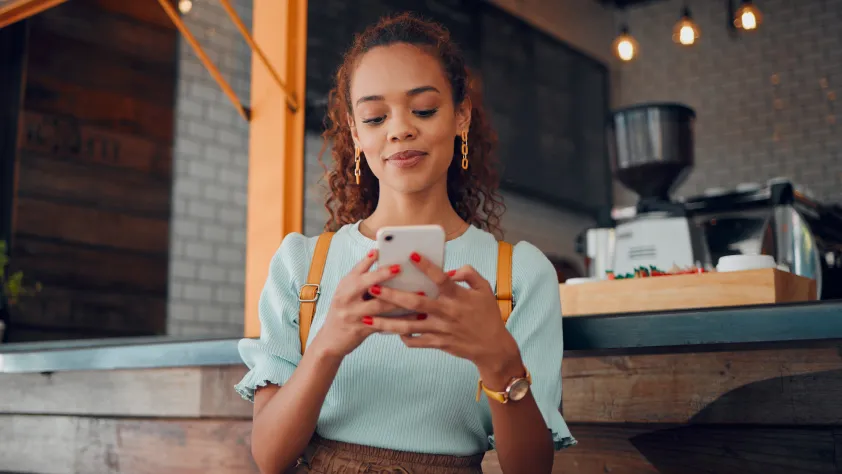 Person standing in cafe making a purchase on their phone through WhatsApp. They are wearing a teal shirt and yellow backpack.