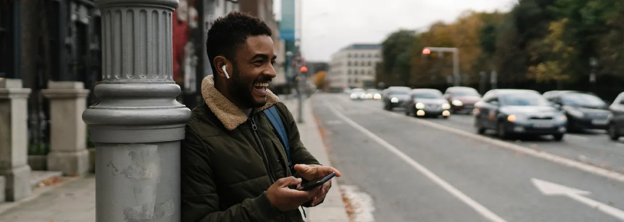 Person wearing a dark green jacket while leaning against a light post on the street. They are laughing and they are wearing AirPods and holding their phone.