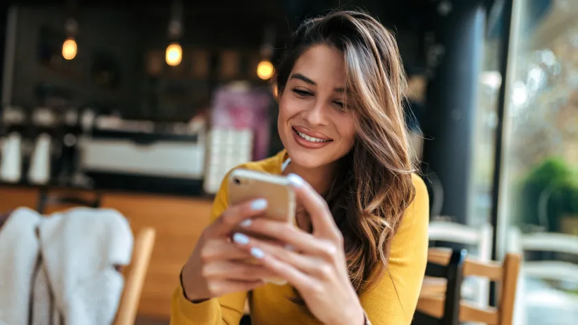 Person sitting in cafe wearing a yellow shirt. They are smiling and talking to a support team via WhatsApp on iPhone.