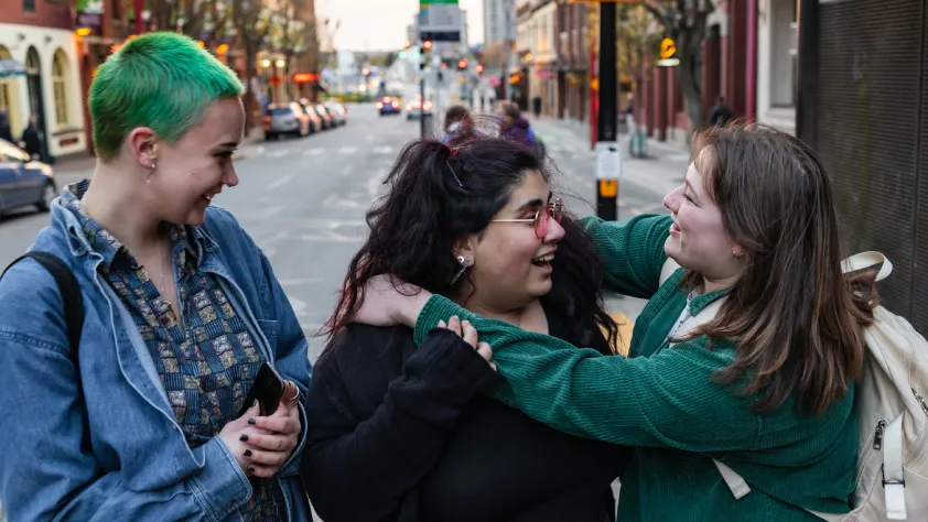 Three friends standing on the sidewalk with the city in the background. They are smiling and two of them are embracing.
