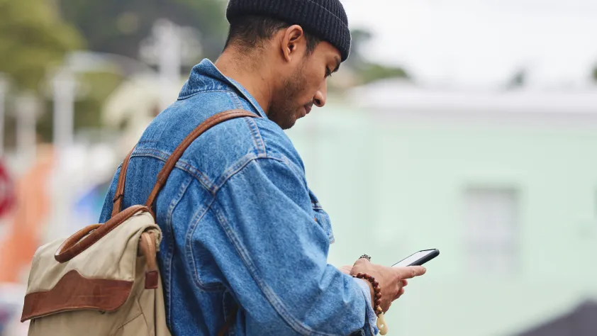 Person walking down the street wearing a jean jacket, black beanie, and backpack. They are looking at their phone and reading a WhatsApp marketing message.