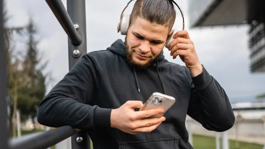Person leaning against outdoor gym equipment. They are wearing headphones and looking at a WhatsApp marketing campaign on their phone.