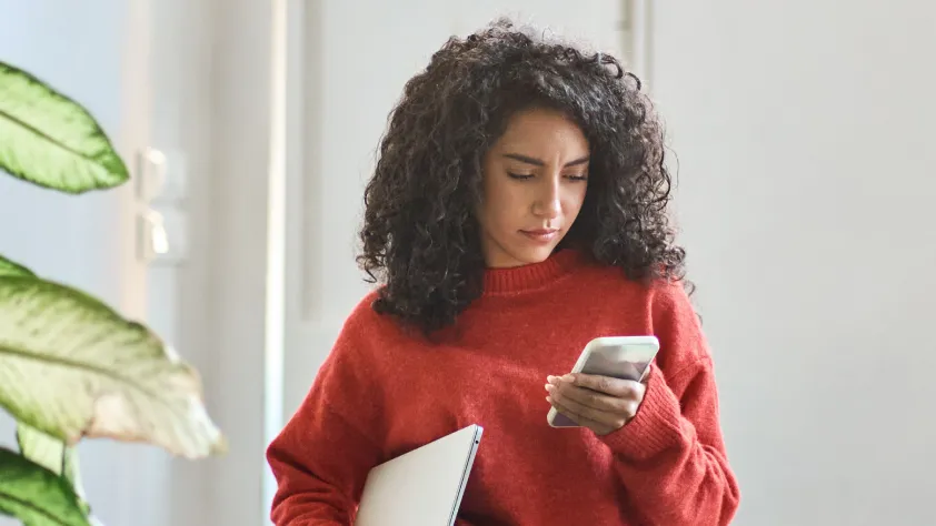 Person walking through office wearing a red sweater. They are carrying a laptop while reading a WhatsApp marketing message on phone.