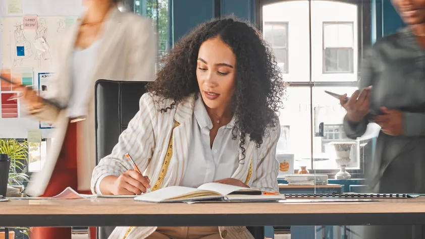 Person sitting at desk and taking notes while two colleagues walk behind them