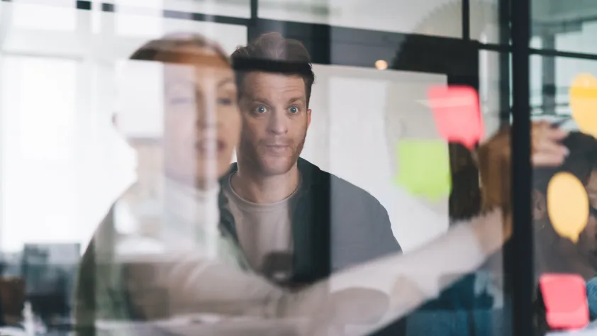 A few people in a conference room during a brainstorming session. One person is writing an idea on the window while another person is watching with their eyes open wide.