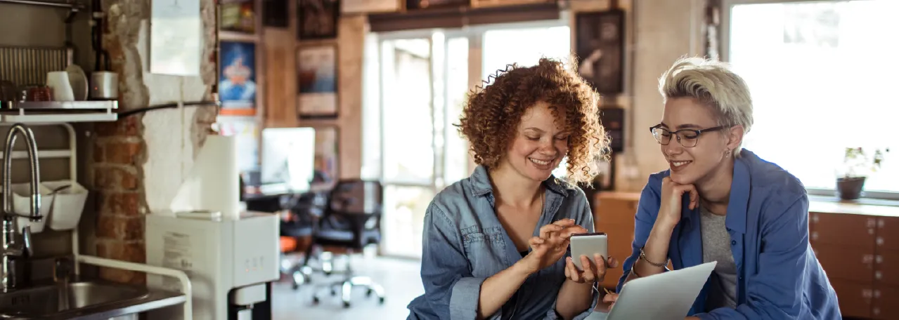 Two people sitting in office kitchen while smiling and looking at phone