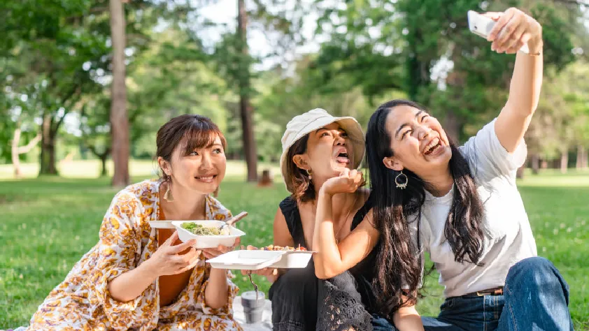 Three friends sitting in a park while eating food and posing for a picture