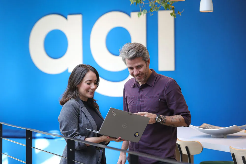 Two individuals standing in front of an Agibank logo sign, both focused on a laptop.