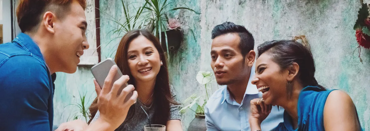 Four people sitting around a lunch table. One person is showing their friends a WhatsApp message on their phone screen.