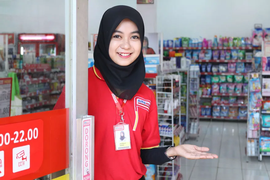 Alfamart staff smiling and gesturing towards entrance to welcome customers into the store
