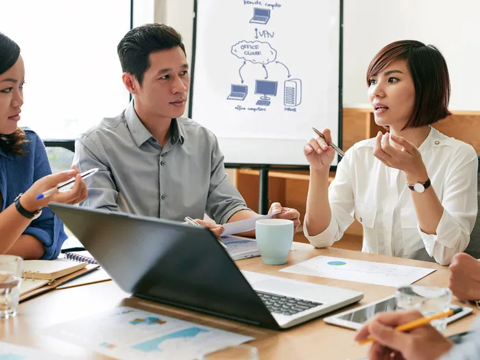 Four coworkers engaged in a meeting, seated at a table with laptops, papers, and cups of coffee, in front of a presentation board displaying a cloud computing diagram