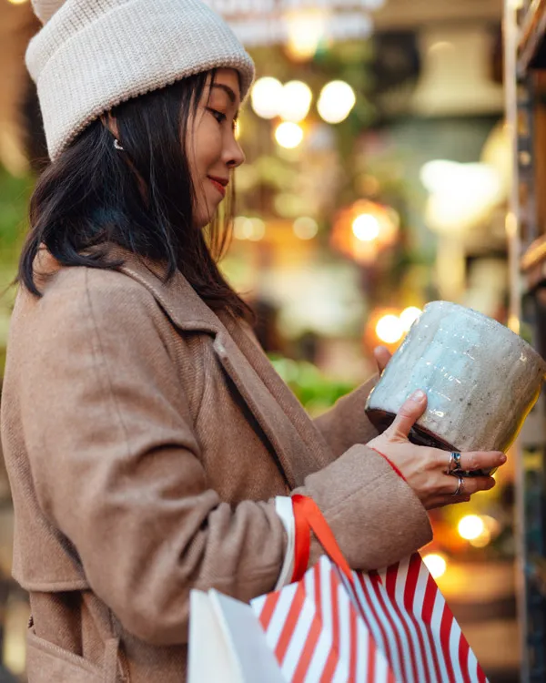 Una mujer vestida con ropa de invierno sonríe mientras sostiene una taza de cerámica y bolsas de compras