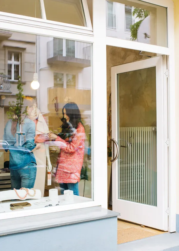Une femme en manteau rouge regarde la vitrine d’un magasin. Son reflet apparaît sur la vitre
