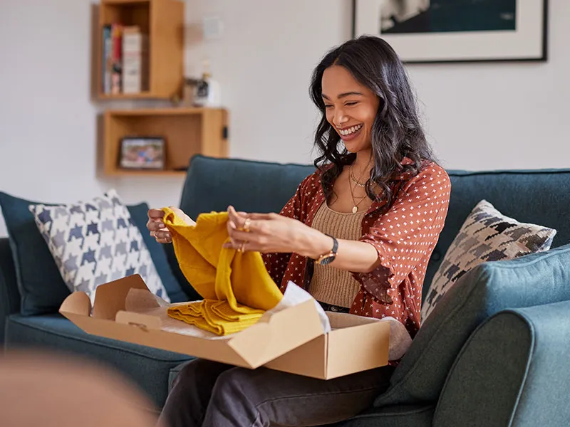 A happy woman sitting on a couch, unwrapping a gift to reveal a yellow sweater.