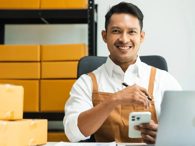 Propriétaire d’entreprise souriant à la caméra, pendant qu’il travaille sur son téléphone et son ordinateur
