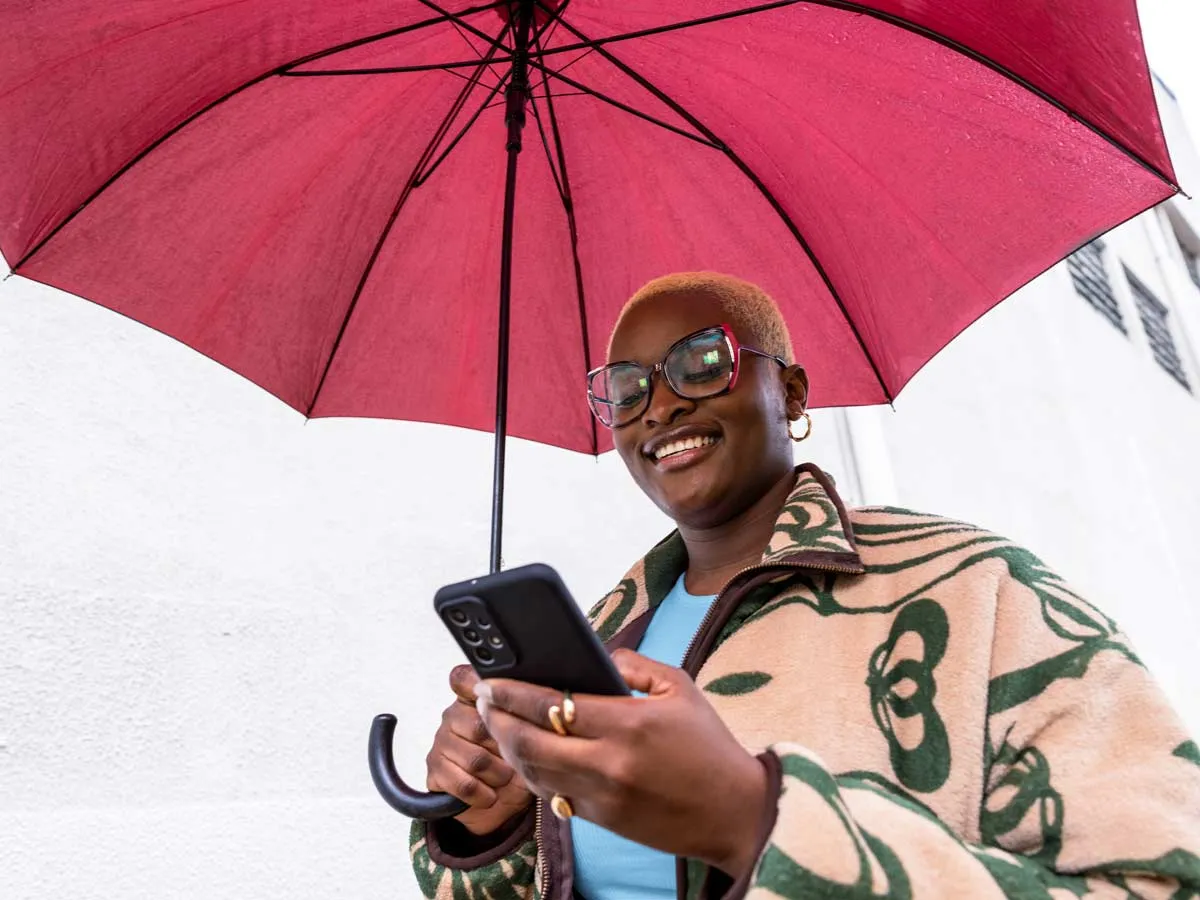 Una mujer sosteniendo un paraguas rojo, caminando afuera y sonriendo mientras mira su teléfono