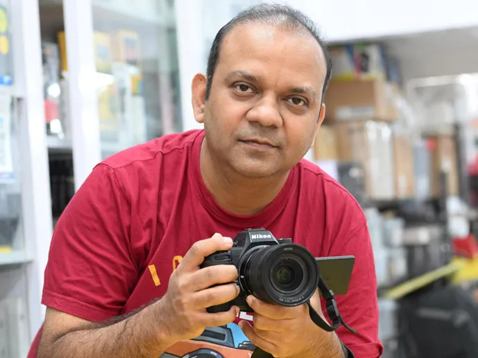 अपने स्टोर में कैमरा को हाथों में पकड़कर सामने की ओर देख रहे एक आदमी की पोट्रेट फ़ोटो