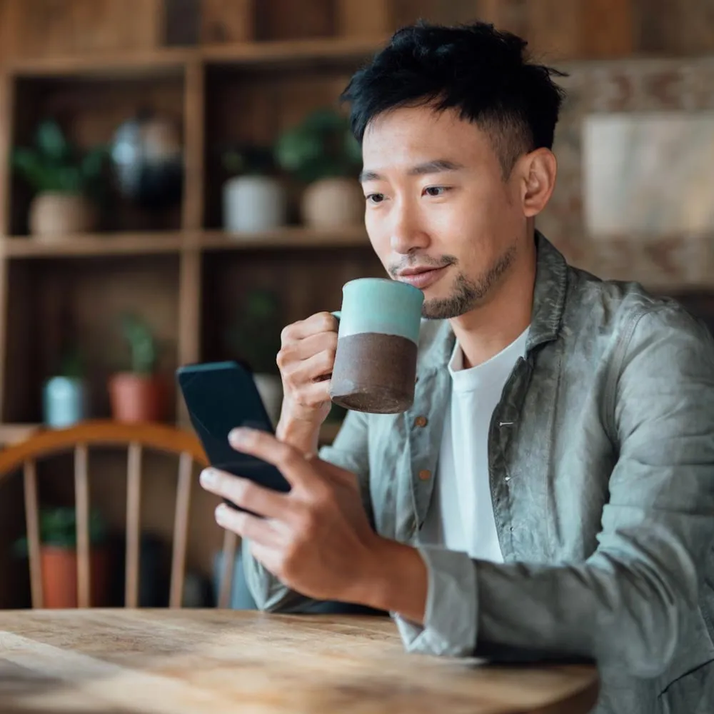 Pria duduk di meja minum dari cangkir kopi dan memakai teleponnya