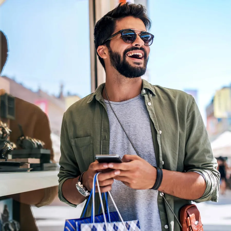 Un hombre sonríe afuera con una bolsa de compras y usa su teléfono